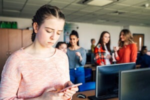 Une adolescente reagit de marniere triste en consultant son smartphone ; en arriere-plan ses camarades de classe semblent se moquer d'elle.