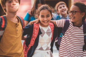 3 enfants bras dessus bras dessous dans la cour de l'ecole avec leur sac a dos.